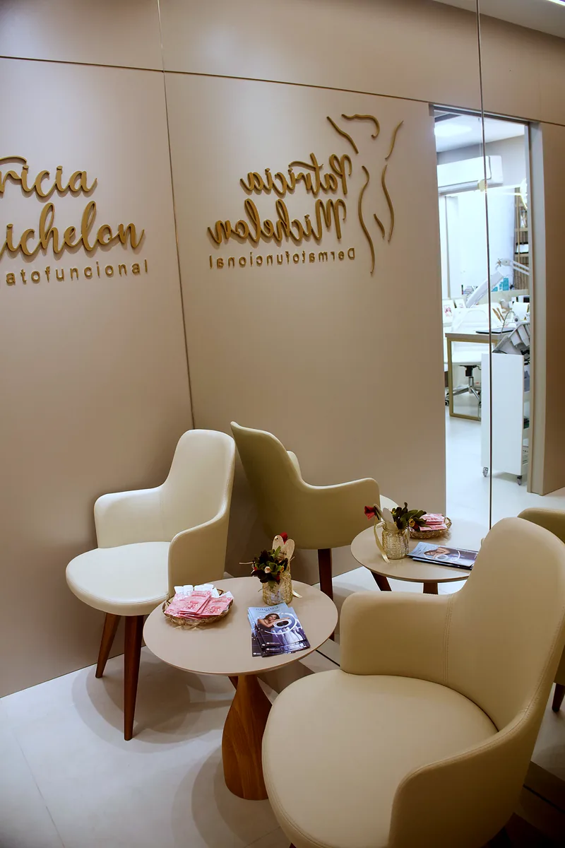 Recepção da clínica com poltronas creme, mesa redonda com flores e o logotipo dourado na parede.